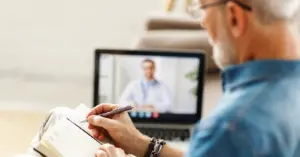 Mental health billing for telehealth services - A senior man taking down notes during an online consultation with his psychologist.