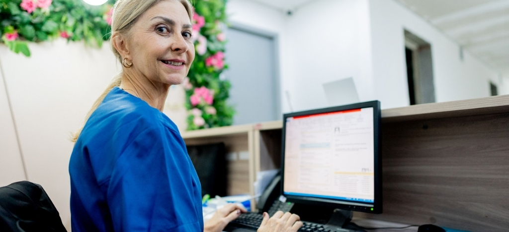 Female front desk medical office staff member working at desktop PC smiles, looking over her shoulder.