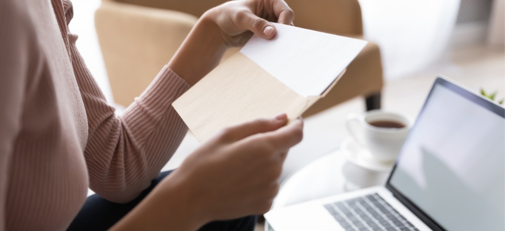 Managing accounts receivable in medical billing - A woman at home opening her mailed medical bill while in front of her laptop.