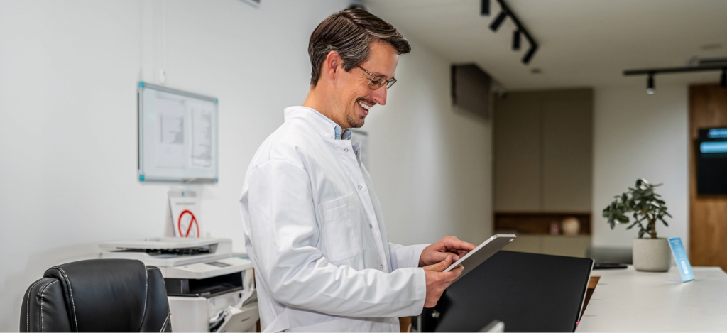 What is practice management software - A smiling male doctor in a hospital using a tablet device to review their practice’s operational workflows.