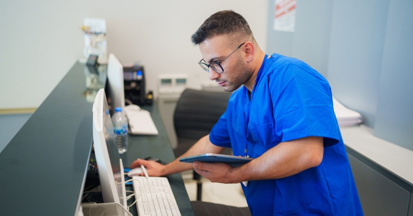 Front desk medical office staff checks the digital tablet in his hand and the reception’s desktop PC.