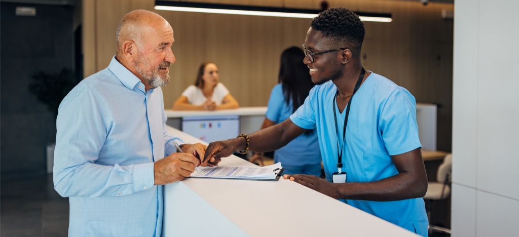 Improving patient financial experience - A male medical professional assisting a senior male patient in signing a form.