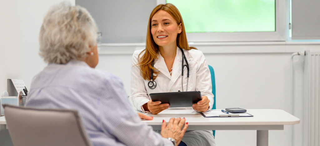 A female doctor holding a tablet device while assisting her senior patient during her consultation.