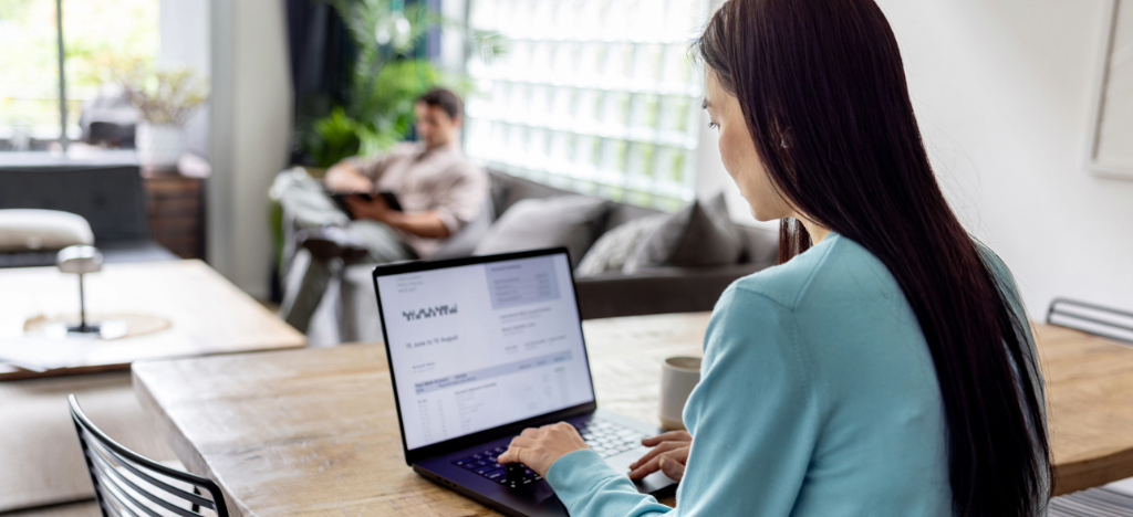 A woman at home with her partner reviewing their online medical bills using a laptop and a tablet device.