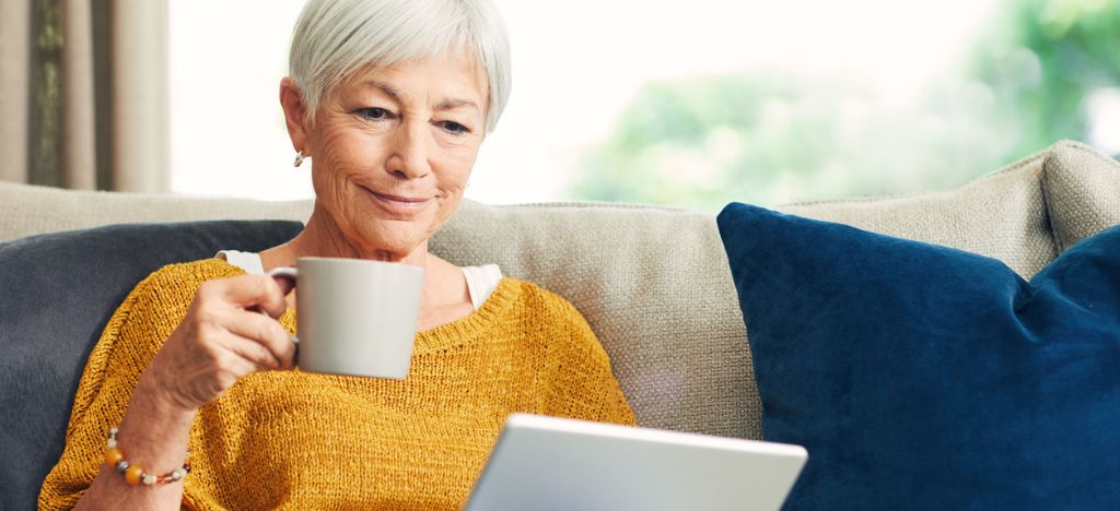 Patient Payment Experience: A senior woman using a digital tablet and having coffee on the sofa at home.